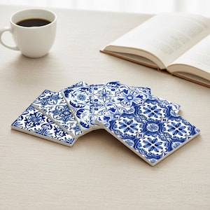 May include: A set of six square ceramic coasters with a blue and white floral pattern. The coasters are arranged in a fan shape on a beige surface, next to a white coffee cup and an open book.