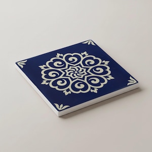 May include: Square ceramic coaster with a navy blue background and a cream-colored floral design. The design is centered and features a symmetrical pattern of swirling shapes. The coaster has a white border and small corner accents.