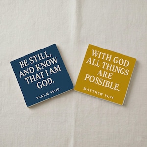 Könnte beinhalten: Zwei quadratische Untersetzer mit inspirierenden Zitaten. Einer ist marineblau mit dem Text "BE STILL, AND KNOW THAT I AM GOD. PSALM 46:10." Der andere ist goldfarben mit dem Text "WITH GOD ALL THINGS ARE POSSIBLE. MATTHEW 19:26."