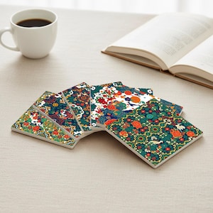 Könnte beinhalten: Eine Sammlung quadratischer Keramikuntersetzer mit farbenfrohen Blumenmustern in Grün, Rot, Orange und Blau. Eine weiße Kaffeetasse und ein offenes Buch befinden sich im Hintergrund.