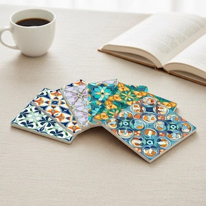 Op de afbeelding: Een verzameling van zes vierkante keramische onderzetters met kleurrijke, patroonontwerpen. De onderzetters hebben verschillende geometrische en bloemmotieven in tinten blauw, oranje, groen en paars. Een witte koffiekop en een open boek zijn op de achtergrond.