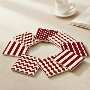 Può includere: Un set di otto sottobicchieri quadrati con vari motivi bordeaux e crema. I disegni includono strisce, zig-zag, pois e forme geometriche. Una tazza e un piattino bianchi sono sullo sfondo.