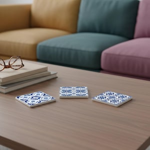 May include: Three square ceramic coasters with a blue and white floral pattern are arranged on a wooden table. The coasters have a glossy finish and a decorative design. The background shows a colorful sofa.