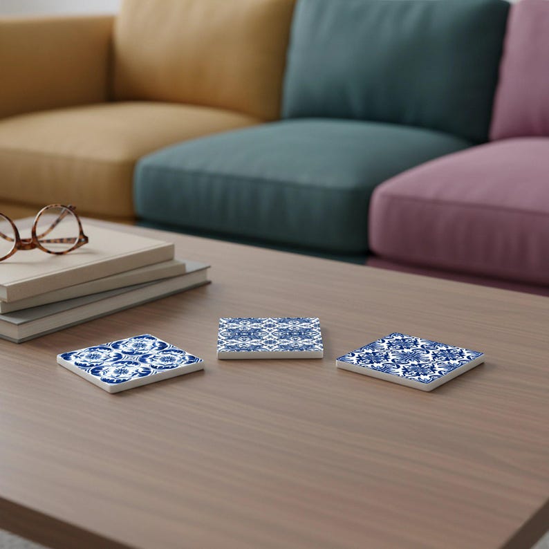 May include: Three square ceramic coasters with a blue and white floral pattern are arranged on a wooden table. The coasters have a glossy finish and a decorative design. The background shows a colorful sofa.