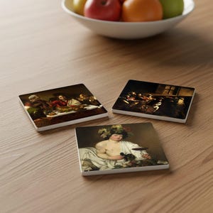 May include: Three square ceramic coasters featuring classic art prints. The coasters depict paintings of figures at a table, a group of people in a dark room, and a person with a wreath. The coasters are on a wooden surface.