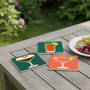 Op de afbeelding: Set van drie vierkante onderzetters met cocktailillustraties. Eén onderzetter toont een bruine cocktail met koffiebonen, een andere een rode cocktail met een limoen en de laatste een rode cocktail met een schijfje citroen. De onderzetters liggen op een houten tafel.
