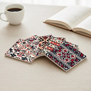May include: A set of six square ceramic coasters with a floral pattern in red, navy, and white. The coasters are arranged in a fan shape on a neutral surface, with a white coffee cup and an open book in the background.