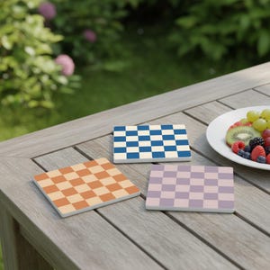 May include: Three rectangular coasters with a checkerboard pattern in orange, blue, and lavender. The coasters are on a wooden table, with a plate of fruit in the background. The coasters are a decorative item.
