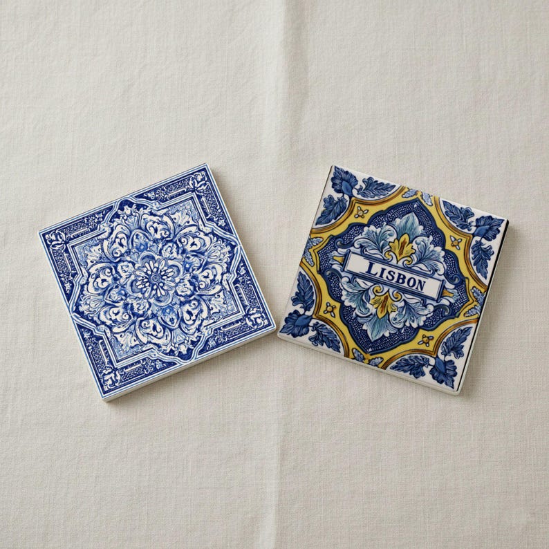 K&ouml;nnte beinhalten: Zwei quadratische Keramikuntersetzer mit aufwendigen blau-wei&szlig;en Blumenmustern. Ein Untersetzer zeigt das Wort "LISBON" in einem rechteckigen Rahmen. Die Untersetzer liegen auf einer wei&szlig;en Oberfl&auml;che.