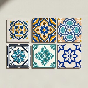 Op de afbeelding: Een set van zes vierkante keramische tegels gerangschikt in twee rijen van drie. Elke tegel heeft een apart geometrisch en bloemmotief in verschillende kleurencombinaties, waaronder blauw, geel, groenblauw, wit en bruin voor een gevarieerd beeld.
