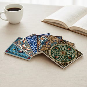 Op de afbeelding: Een verzameling van zes vierkante keramische onderzetters met ingewikkelde, kleurrijke patronen. De onderzetters hebben ontwerpen in blauw, oranje, groen en goud. Een witte koffiekop en een open boek liggen ook op de tafel.