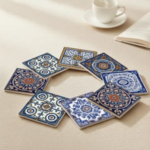 Könnte beinhalten: Acht quadratische Keramikuntersetzer mit farbenfrohen Mustern. Die Untersetzer zeigen blaue, weiße, orangefarbene und goldene Designs, angeordnet in einem Kreis. Im Hintergrund sind eine weiße Tasse und Untertasse zu sehen.