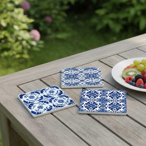 May include: Three square ceramic coasters with blue and white floral patterns are arranged on a wooden table. The coasters have a glossy finish and are surrounded by a blurred background of greenery and a plate of fruit.