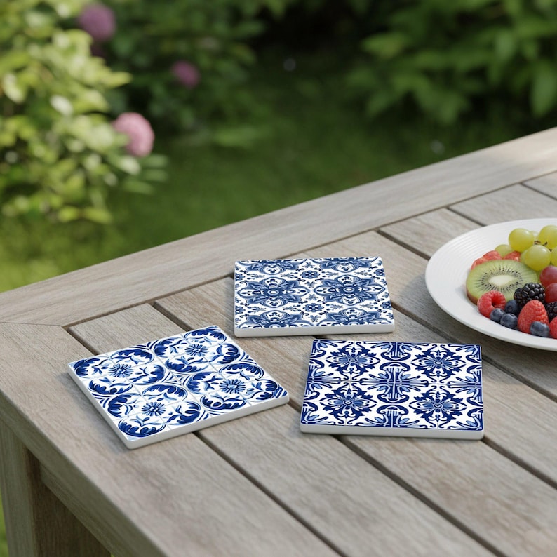 May include: Three square ceramic coasters with blue and white floral patterns are arranged on a wooden table. The coasters have a glossy finish and are surrounded by a blurred background of greenery and a plate of fruit.