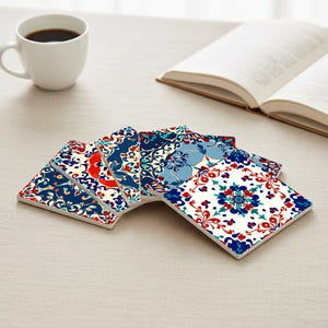 May include: A collection of six square ceramic coasters featuring detailed floral designs in blue, red, and white. The coasters are displayed on a neutral surface, with a coffee cup and an open book visible.