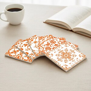 Könnte beinhalten: Ein Satz von fünf quadratischen Untersetzern mit weißem Hintergrund und einem orangefarbenen Blumenmuster. Eine weiße Kaffeetasse mit dunkler Flüssigkeit und ein offenes Buch befinden sich im Hintergrund. Die Untersetzer sind fächerförmig angeordnet.