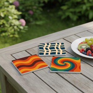 Könnte beinhalten: Drei quadratische Keramikuntersetzer mit Retro-Designs liegen auf einem Holztisch. Ein Untersetzer zeigt orangefarbene und braune Wellenlinien, ein anderer einen Wirbel aus Grün, Orange und Gelb, und der dritte ein blaues und beiges Muster.