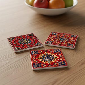 May include: Three square coasters with a red background and an intricate, ornate design in gold and navy blue. The coasters are arranged on a light brown wooden surface, with a bowl of fruit in the background.