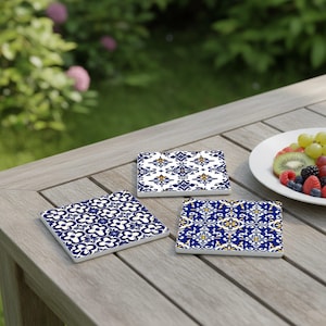 May include: Three square ceramic coasters with blue and white floral patterns are arranged on a wooden table. The coasters have different designs, with one featuring a repeating flower pattern and the others showcasing intricate geometric motifs.