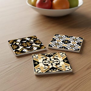 May include: Three square coasters with geometric designs in black, gold, and white are arranged on a wooden surface. The coasters feature a repeating pattern of starburst and angular shapes. A bowl of fruit sits in the background.