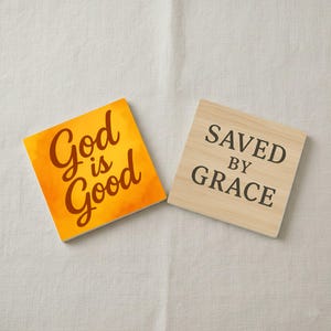 Könnte beinhalten: Zwei quadratische Untersetzer. Einer hat den Text "God is Good" in brauner Schrift auf orangefarbenem Hintergrund. Der andere hat den Text "Saved by Grace" in schwarzer Schrift auf hellem Holzmaserungshintergrund.