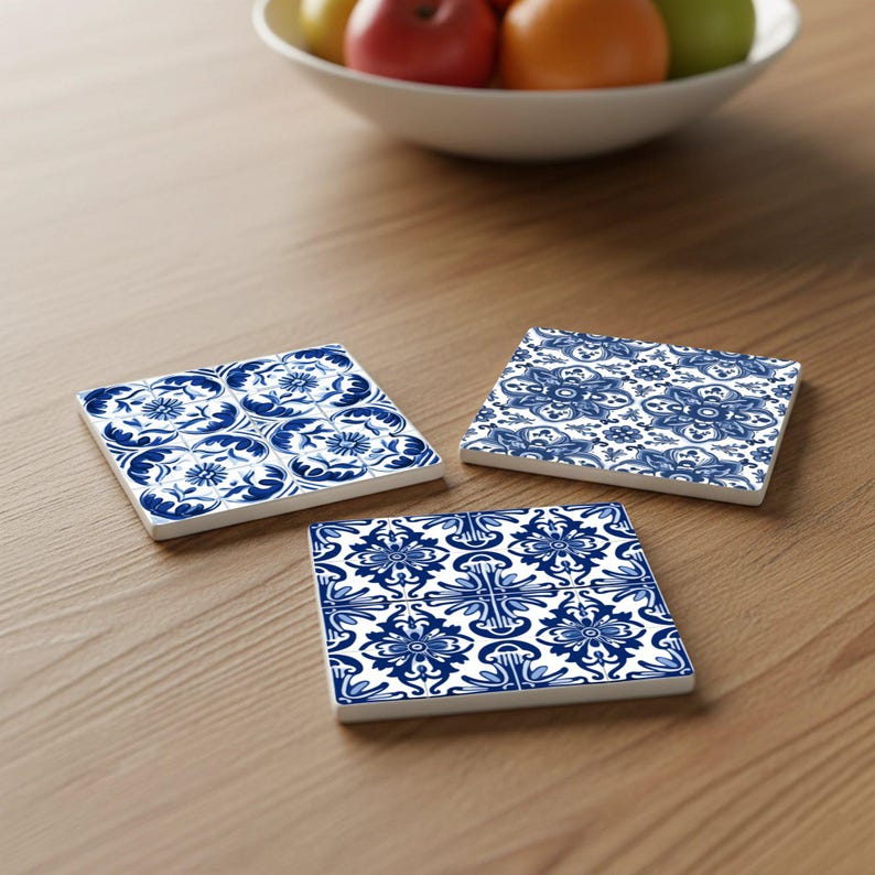 May include: Three square ceramic coasters with blue and white floral patterns are arranged on a wooden surface. The coasters feature different designs, including stylized flowers and geometric shapes. A bowl of fruit sits in the background.