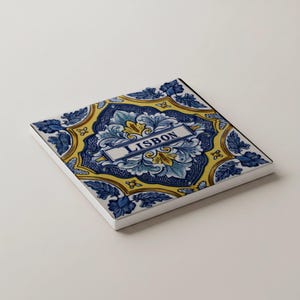 K&ouml;nnte beinhalten: Quadratische Keramikfliese mit einem dekorativen Design in Blau, Gelb und Wei&szlig;. Die Fliese zeigt florale Muster und das Wort "LISBON" in einem rechteckigen Banner. Die Fliese hat einen wei&szlig;en Rand.