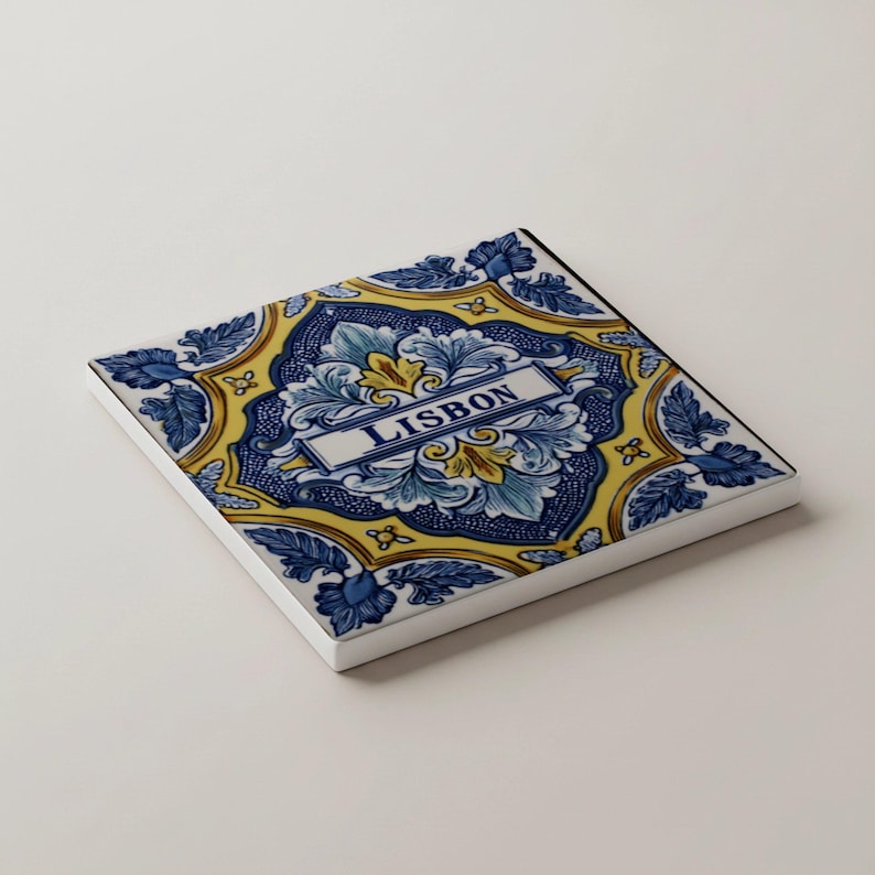 K&ouml;nnte beinhalten: Quadratische Keramikfliese mit einem dekorativen Design in Blau, Gelb und Wei&szlig;. Die Fliese zeigt florale Muster und das Wort "LISBON" in einem rechteckigen Banner. Die Fliese hat einen wei&szlig;en Rand.