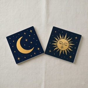 Puede incluir: Dos posavasos cuadrados con diseños celestiales. Uno presenta una luna creciente dorada y estrellas sobre un fondo azul marino. El otro tiene un sol dorado con ojos cerrados y rayos, también sobre un fondo azul marino. Los posavasos son probablemente de cerámica.