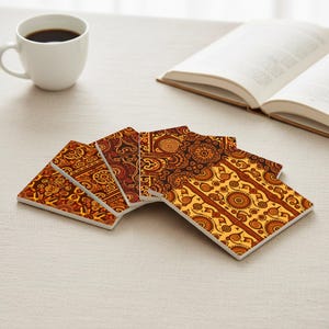 May include: Set of six square ceramic coasters with a repeating, intricate pattern in shades of brown, orange, and yellow. A white coffee mug filled with dark liquid and an open book are in the background.