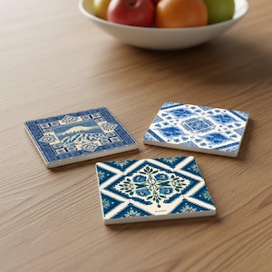 Könnte beinhalten: Drei quadratische Keramikuntersetzer mit blau-weißen Mustern. Einer zeigt eine Berglandschaft, ein anderer ein Blumenmuster und der dritte ein geometrisches Muster. Die Untersetzer liegen auf einer Holzoberfläche.