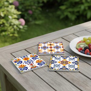 Puede incluir: Tres posavasos cuadrados de cerámica con coloridos diseños florales y geométricos en azul, amarillo y rojo. Los posavasos están sobre una mesa de madera, con un plato de fruta en el fondo.