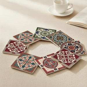 Puede incluir: Un conjunto de posavasos cuadrados de cerámica con intrincados y coloridos patrones geométricos. Los posavasos presentan diseños en tonos rojos, azules, verdes y crema, dispuestos en un patrón circular. Una taza de té blanca y un libro están en el fondo.