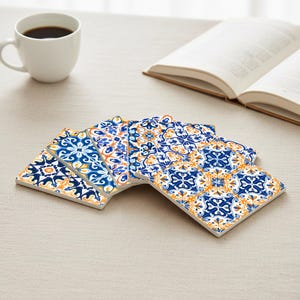 Op de afbeelding: Een stapel keramische onderzetters met een blauw, oranje en wit bloemmotief. Een witte koffiemok met zwarte koffie en een open boek staan op de achtergrond. De onderzetters zijn vierkant met een decoratieve rand.