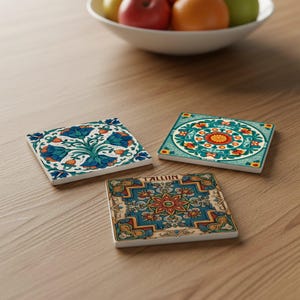 Könnte beinhalten: Drei quadratische Keramikuntersetzer mit farbenfrohen Blumen- und geometrischen Mustern. Die Untersetzer zeigen Designs in Türkis, Orange und Blau auf weißem Grund. Auf einem Untersetzer steht das Wort "TALLIN".