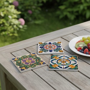 Könnte beinhalten: Drei dekorative Keramikuntersetzer mit farbenfrohen floralen Designs. Die Untersetzer haben eine quadratische Form und liegen auf einem Holztisch. Die Designs umfassen Blau-, Grün-, Gelb-, Lila- und Orangetöne. Ein Teller mit Obst ist ebenfalls zu sehen.