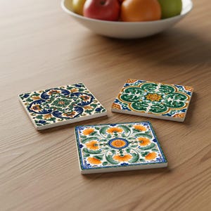 May include: Three square ceramic coasters with colourful floral designs. The coasters feature intricate patterns in shades of blue, green, orange, and yellow, set against a white background. The coasters are displayed on a wooden surface.