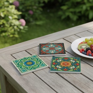 Op de afbeelding: Drie keramische onderzetters met kleurrijke bloemenpatronen liggen op een houten tafel. De onderzetters hebben ingewikkelde patronen in groen, blauw, oranje en rood. Een bord met fruit staat in de buurt. Afmetingen: 10 cm x 10 cm.