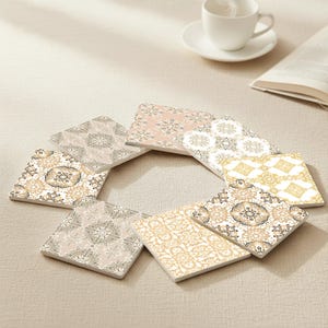 May include: A collection of eight square ceramic coasters arranged in a circle. Each coaster features a unique, intricate floral pattern in shades of beige, gold, and pink. A white teacup and saucer are in the background.
