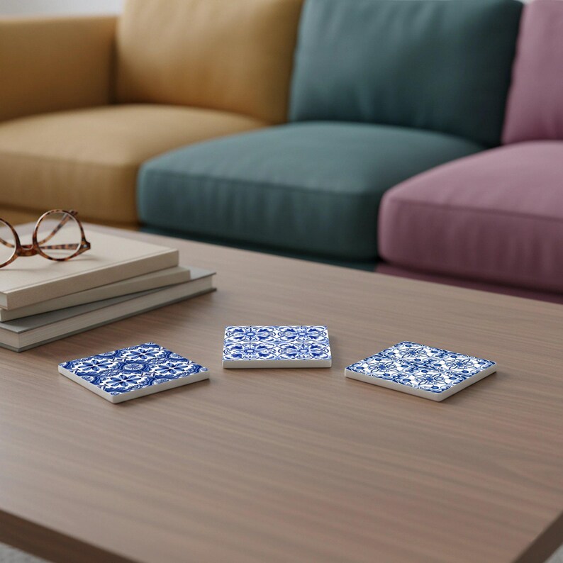 May include: Three square ceramic coasters with a blue and white floral pattern are displayed on a wooden table. The coasters are arranged in a triangular formation, showcasing their decorative design. The background features a sofa.