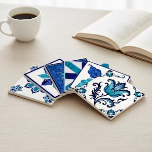 Peut inclure: Ensemble de dessous de verre carrés en céramique avec des motifs floraux et géométriques bleus complexes. Les dessous de verre sont disposés sur une surface neutre, avec une tasse à café blanche et un livre ouvert. Les motifs varient en nuances de bleu.