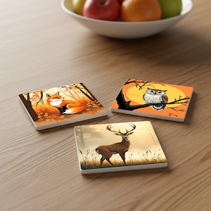 Op de afbeelding: Set van drie vierkante keramische onderzetters met bosdierontwerpen. Eén onderzetter toont een vos in herfstbladeren, een andere een uil op een tak en de derde een hert in een mistig bos. De onderzetters liggen op een houten oppervlak.