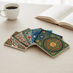 May include: A selection of square ceramic coasters featuring detailed floral and geometric designs in green, blue, red, and gold. The coasters are fanned out on a neutral surface. A white coffee cup and an open book are visible in the background.
