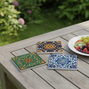 May include: Three square ceramic coasters with intricate floral patterns in shades of blue, green, and yellow. The coasters are arranged on a wooden table, suggesting outdoor use. A plate of fruit is in the background.