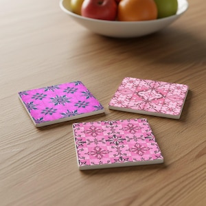 Pode incluir: Três bases de cerâmica quadradas com padrões florais em tons de rosa e roxo. As bases estão dispostas sobre uma superfície de madeira, com uma tigela de frutas ao fundo.