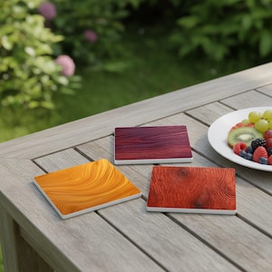 Könnte beinhalten: Drei quadratische Untersetzer mit einzigartigen Holzmaserungsdesigns in Orange-, Rot- und Lilatönen. Die Untersetzer liegen auf einem Holztisch, zusammen mit einem Teller mit frischem Obst.