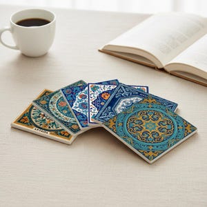 May include: A collection of square ceramic coasters with intricate, colorful patterns. The coasters feature floral and geometric designs in shades of blue, gold, and teal. One coaster has the word "ANTALYA" printed on it. A white coffee cup and an open book are in the background.
