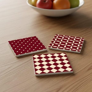 Könnte beinhalten: Drei quadratische Untersetzer mit verschiedenen geometrischen Mustern. Einer hat einen weinroten Hintergrund mit weißen Rauten. Ein anderer hat einen weinroten Hintergrund mit einem weißen geometrischen Design. Der dritte hat einen cremefarbenen Hintergrund mit weinroten Rauten.