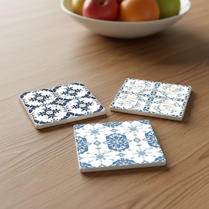 Op de afbeelding: Drie vierkante keramische onderzetters met ingewikkelde blauw-witte bloempatronen. De onderzetters zijn gerangschikt op een houten oppervlak, met een fruitschaal op de achtergrond. Elke onderzetter heeft een unieke ontwerpvariatie.