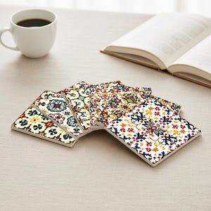 Puede incluir: Un conjunto de posavasos cuadrados de cerámica con intrincados patrones coloridos. Los posavasos presentan diseños en rojo, azul, amarillo y crema, dispuestos en forma de abanico. Una taza de café blanca y un libro abierto están en el fondo.
